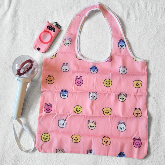 A pink tote bag with Stray Kids's Skzoo characters on a white background. A 1st generation stray kids lightstick is on the left side. The tote storage clip-on bag is on the top-left of the image.