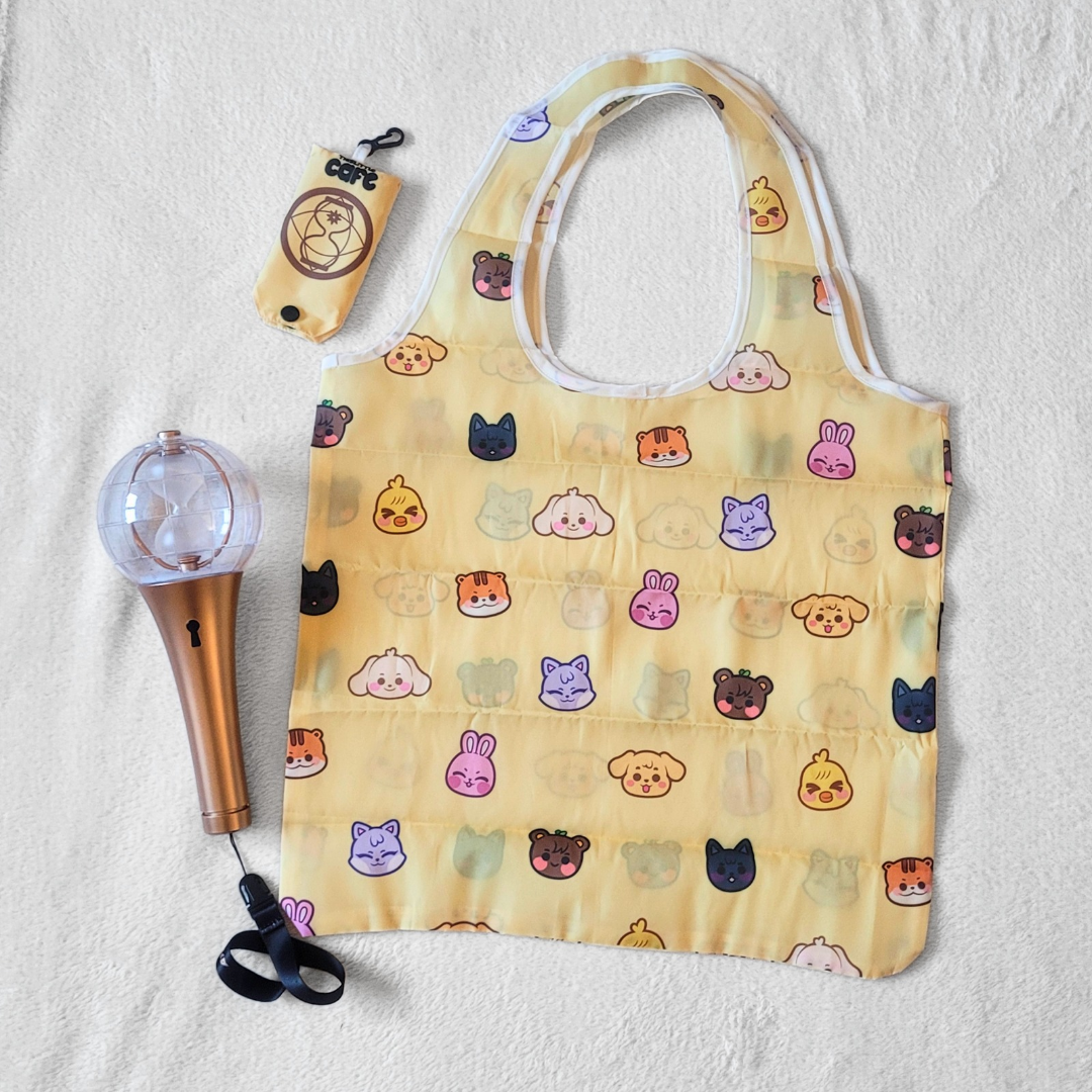 A yellow tote bag with Ateez's Anitee, characters on a white background. A 2nd generation Ateez lightstick is on the left side. The tote storage clip-on bag is on the top-left of the image.
