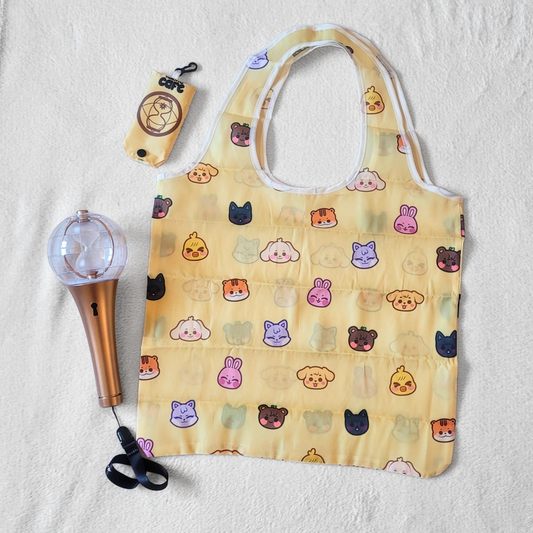 A yellow tote bag with Ateez's Anitee, characters on a white background. A 2nd generation Ateez lightstick is on the left side. The tote storage clip-on bag is on the top-left of the image.